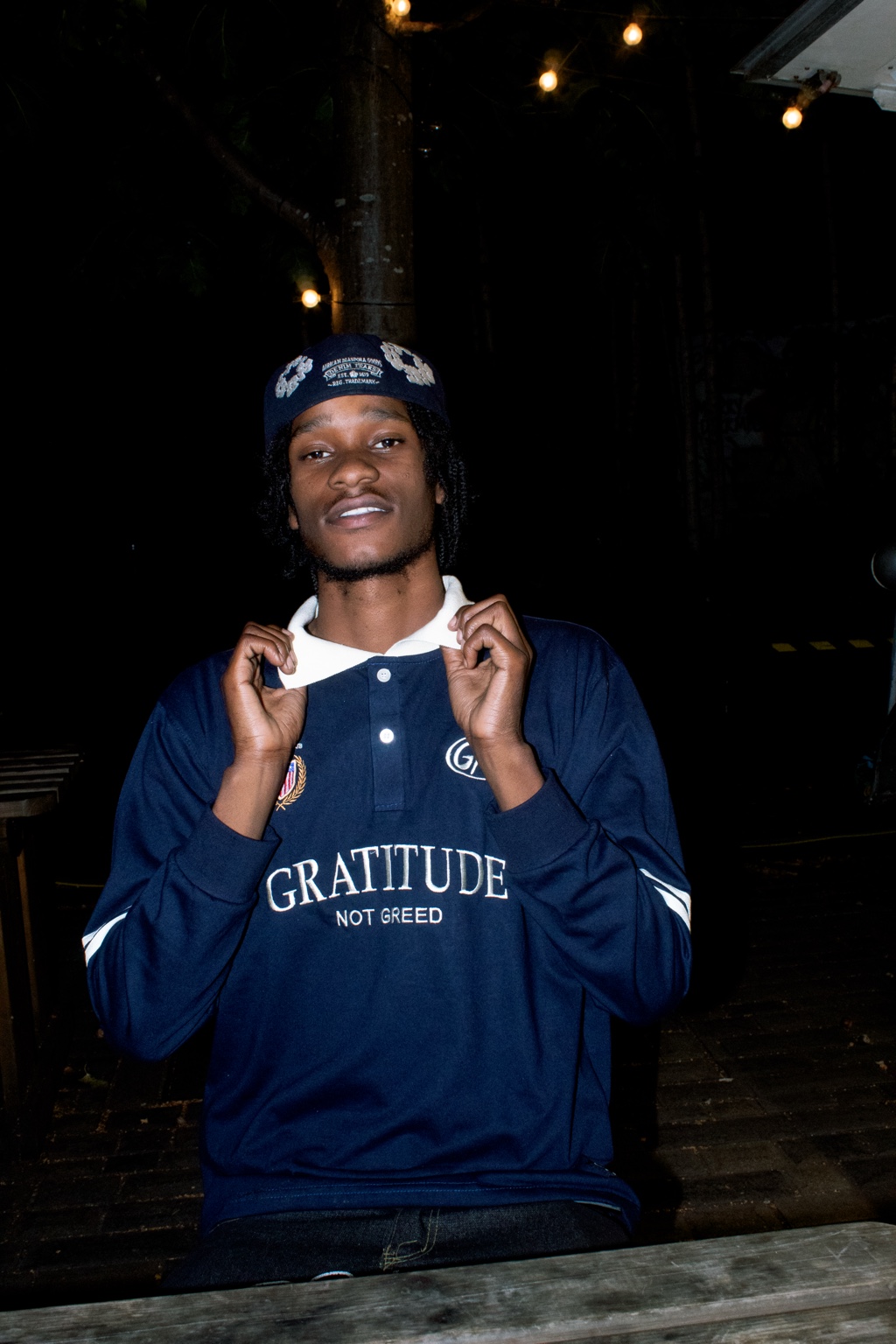 GNG campaign — navy Gratitude Not Greed rugby polo, white collar held up, worn at night under string lights