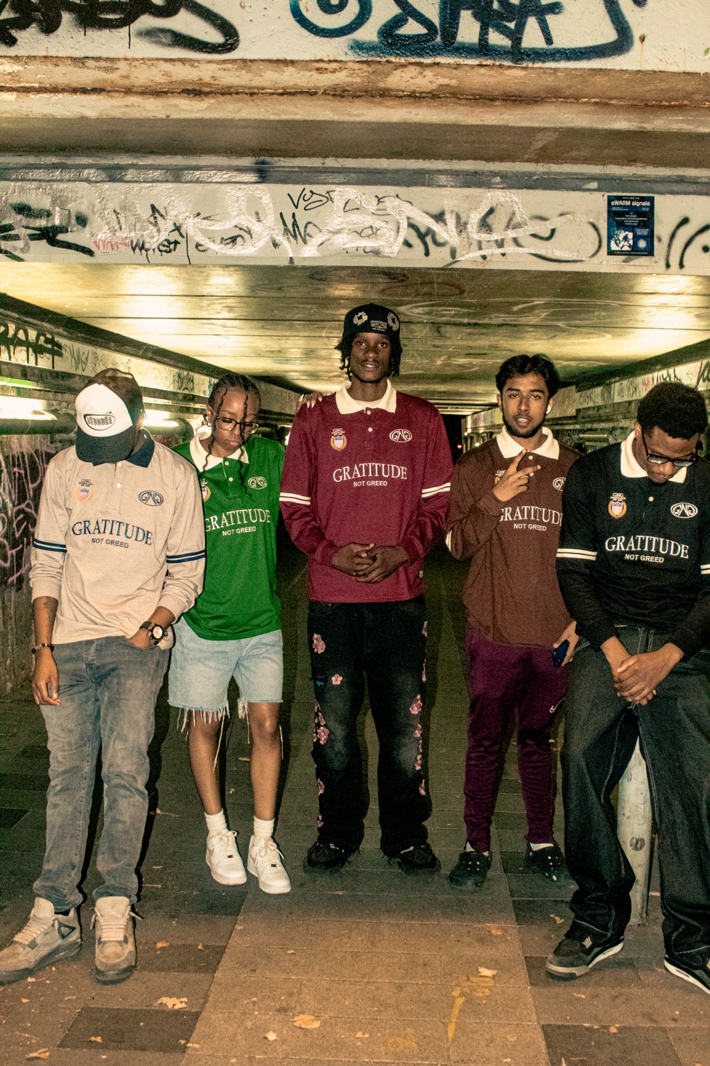 GNG campaign — five people wearing all five Gratitude Not Greed rugby polo colourways under a graffiti-covered London tunnel