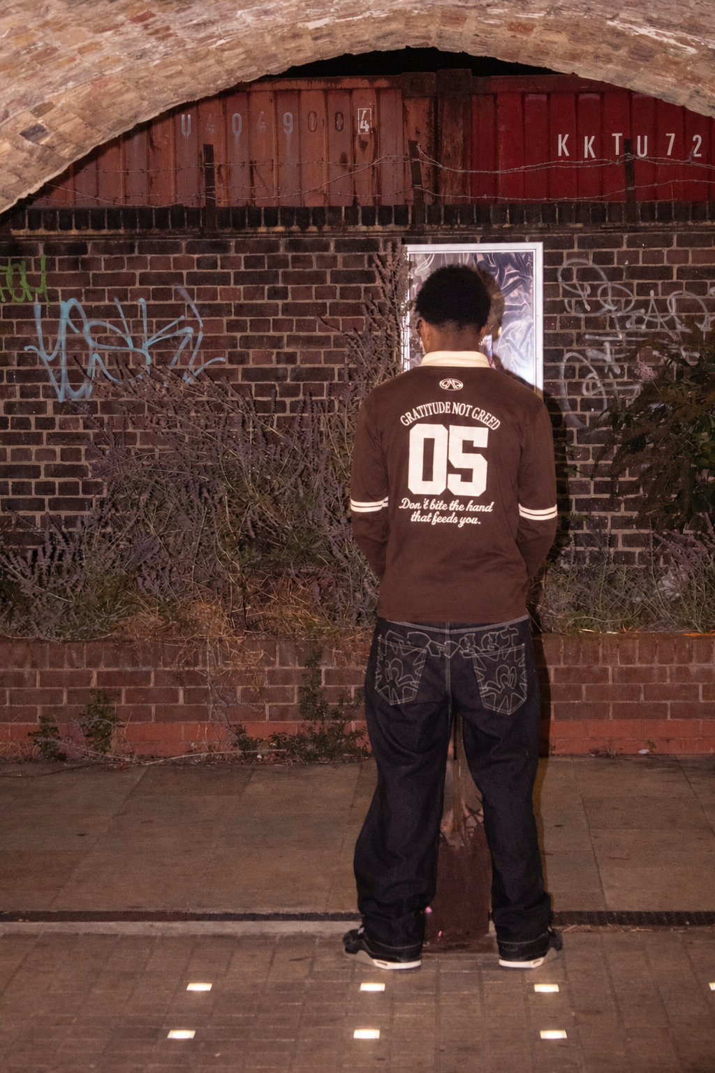 GNG campaign — 05 back of the brown rugby polo, Don't bite the hand that feeds you embroidery, under a graffitied London railway arch at night